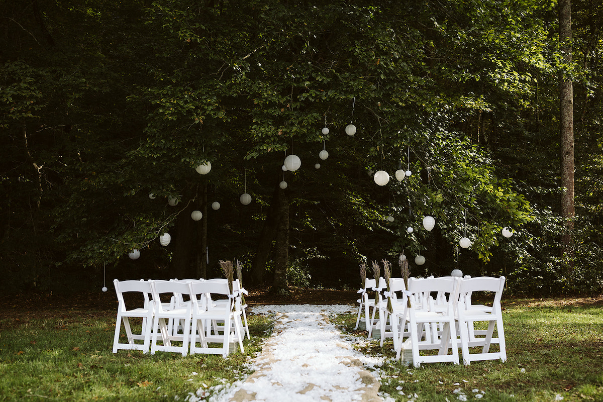 a small number of white folding chairs sits on a lawn on either side of a white flower petal-lined aisle