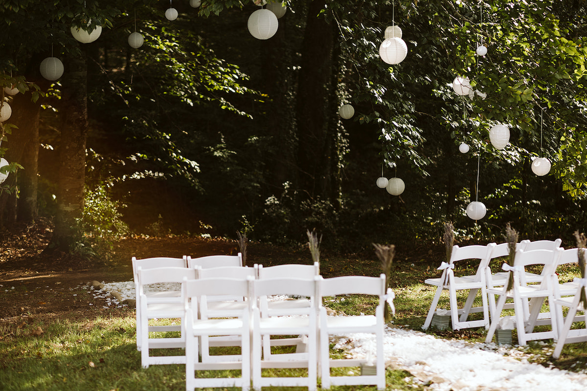 white paper lanterns hang from large trees on the edge of a field and woods. white folding chairs are arranged nearby