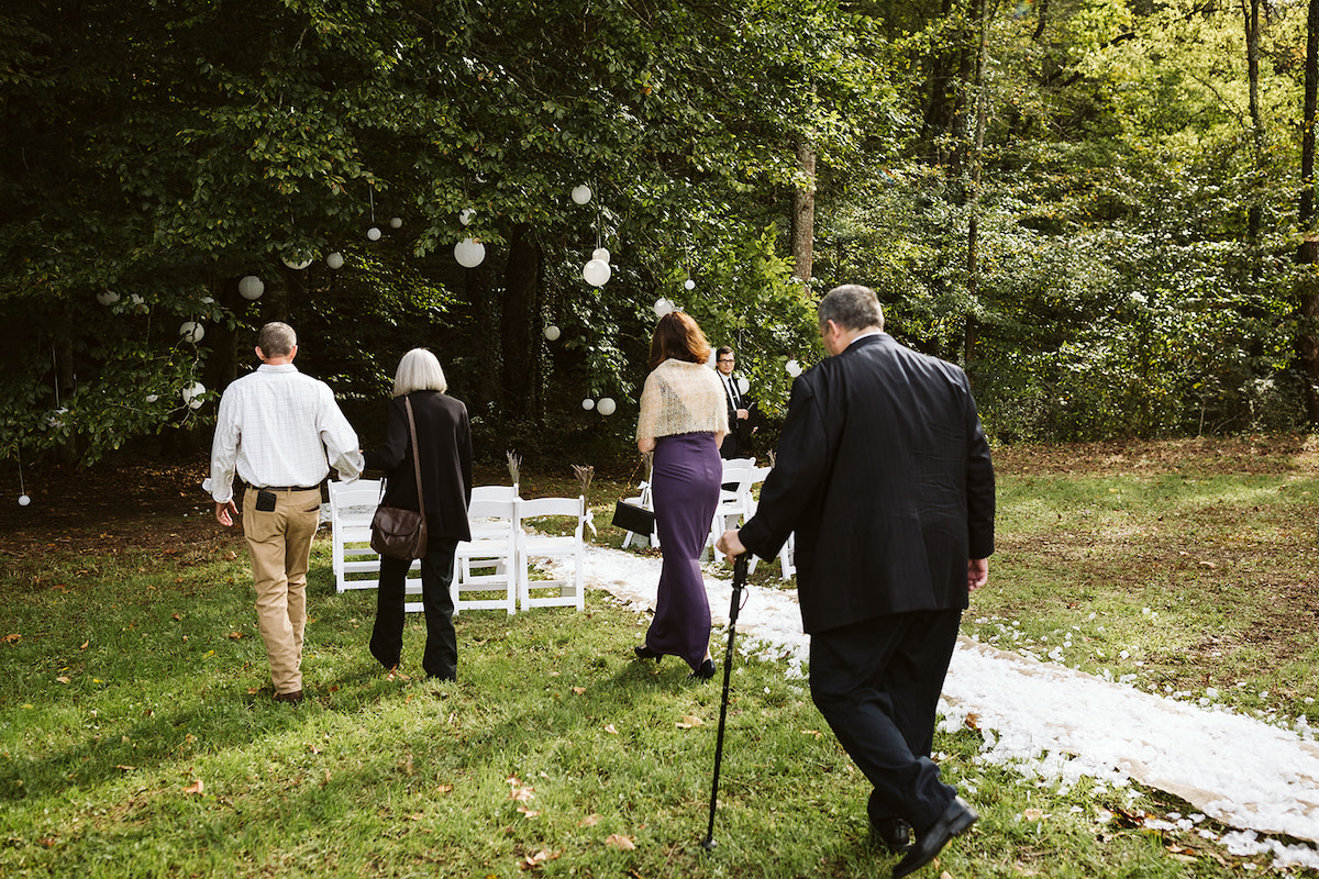 people walk in the grass toward folding white chairs set up at the edge of lawn and woods