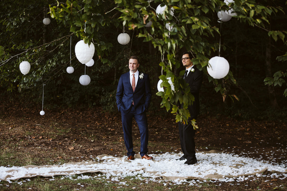groom stands under low branches of a tree, surrounded by white paper lanterns. The ground is covered with white petals