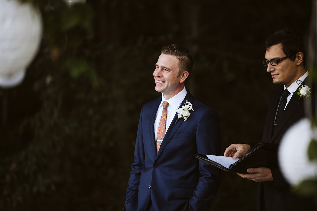 groom in deep blue suit and white boutonniere smiles as he stands next to officiant