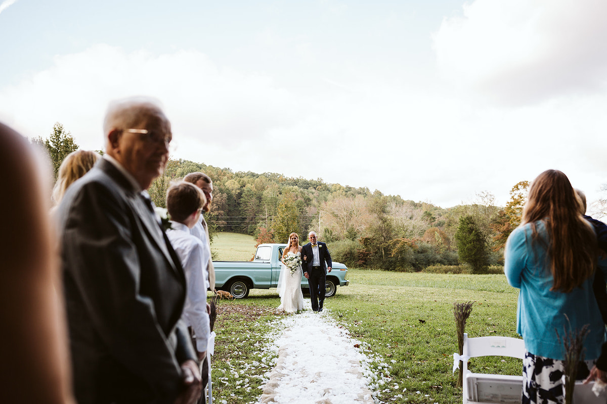 bride and her father walk down a path of white flower petals through a grassy field toward wedding guests