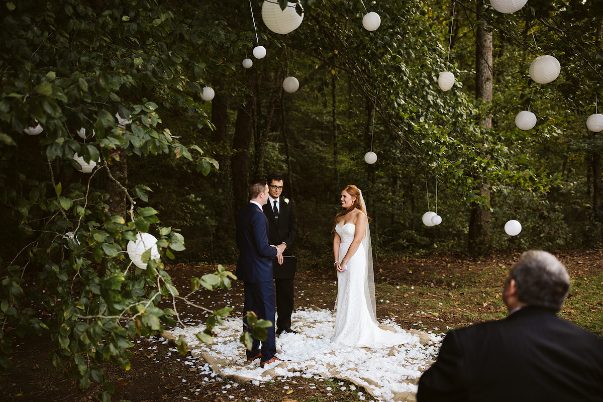bride smiles as groom reads his vows. They are surrounded by trees and white paper lanterns and stand on bed of white petals