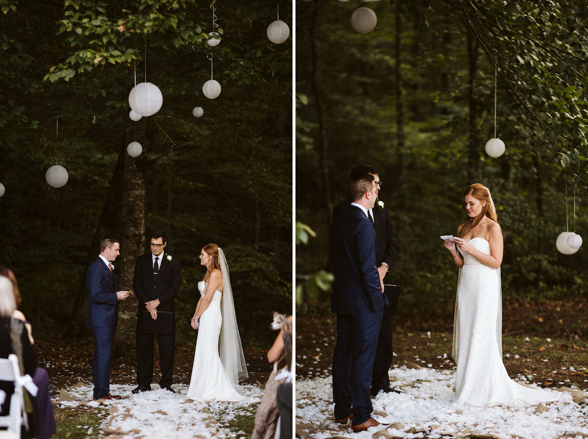 bride smiles as groom reads his vows. They are surrounded by trees and white paper lanterns and stand on bed of white petals