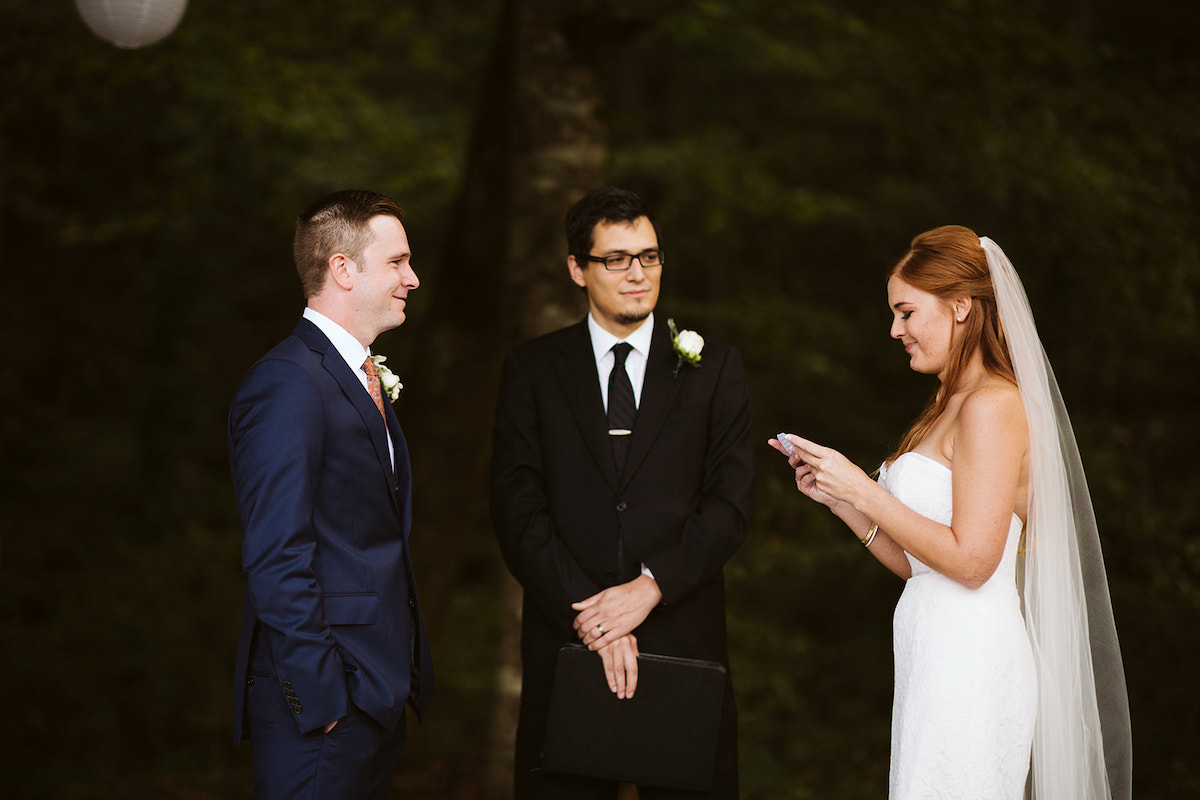 groom looks at bride as she reads vows. They are surrounded by trees and paper lanterns and stand on bed of white petals