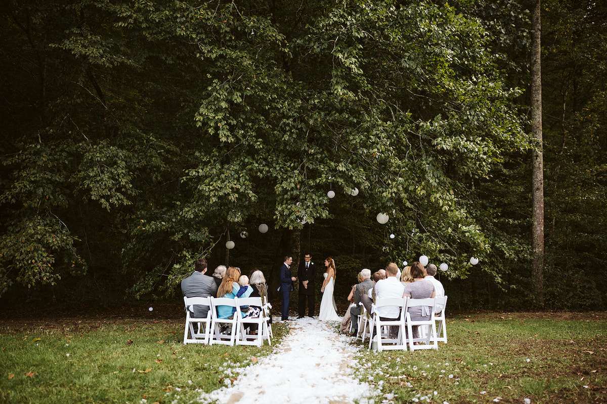 groom looks at bride as she reads vows. They are surrounded by trees and paper lanterns and stand on bed of white petals