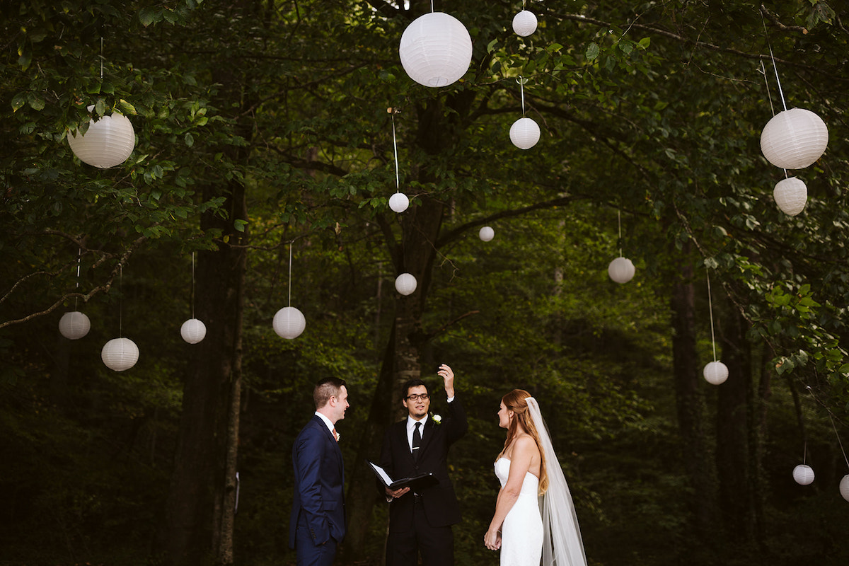 bride and groom stand before officiant. They are surrounded by tall green trees and white paper lanterns