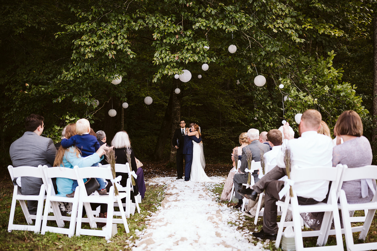 Bride and groom hug beneath a large tree strung with white paper lanterns. Wedding guests watch from white folding chairs.