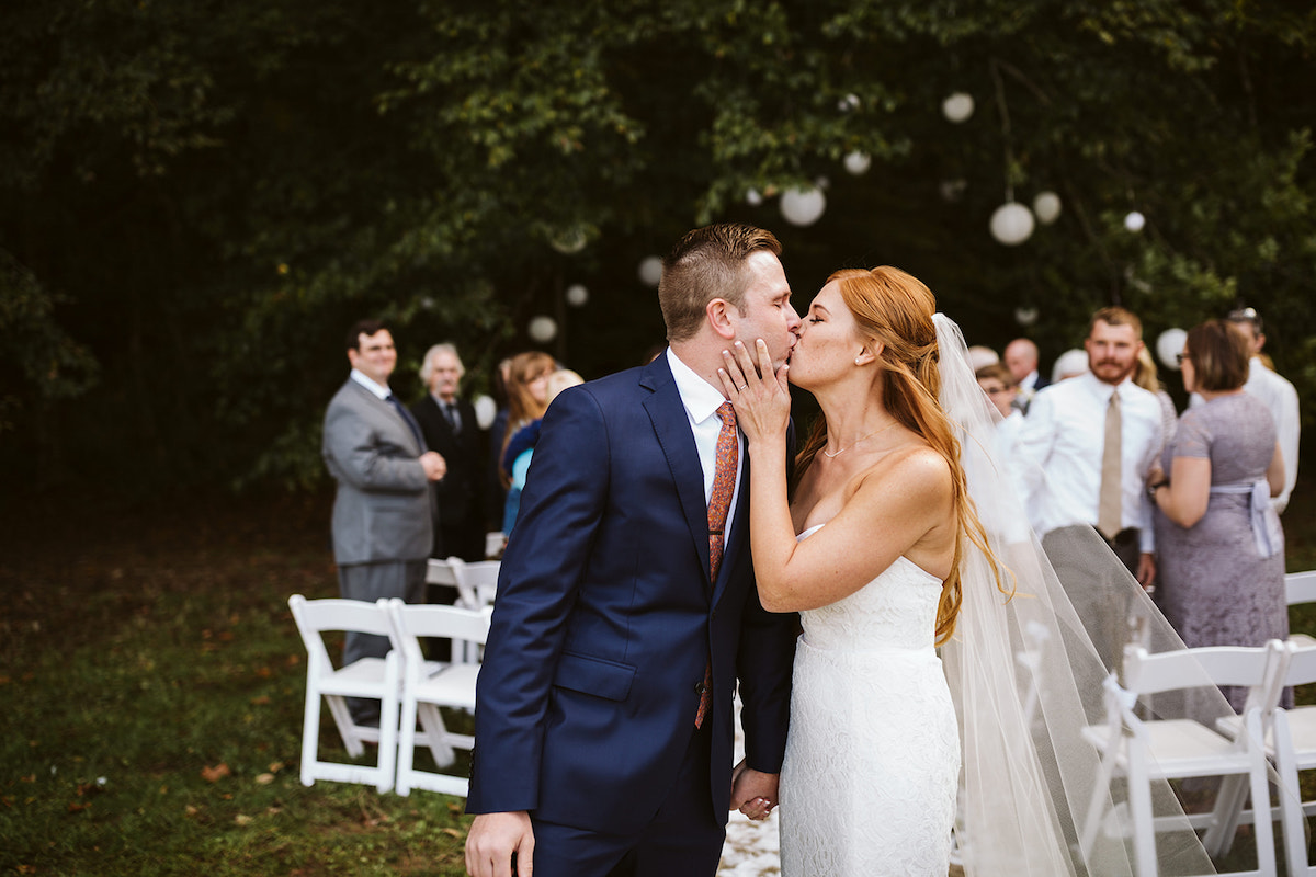 bride in strapless dress and groom in dark blue suit kiss as wedding guests look on