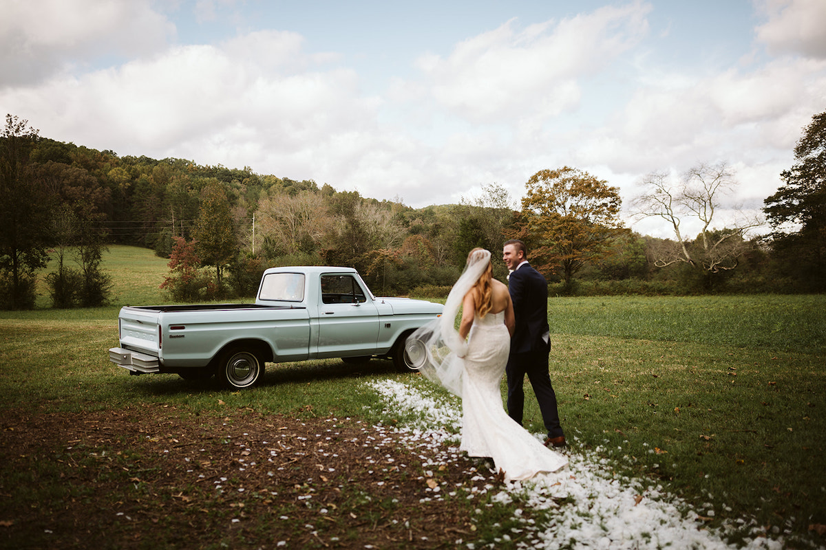 bride and groom walk on a bed of white flower petals toward antique baby blue Ford F-100 pickup truck