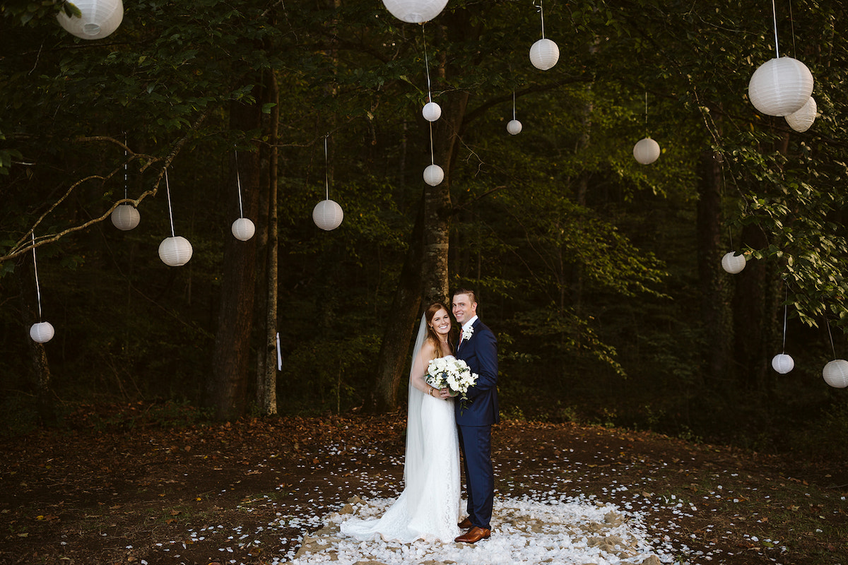 bride and groom stand on a bed of white petals beneath tall green trees hung with white paper lanterns