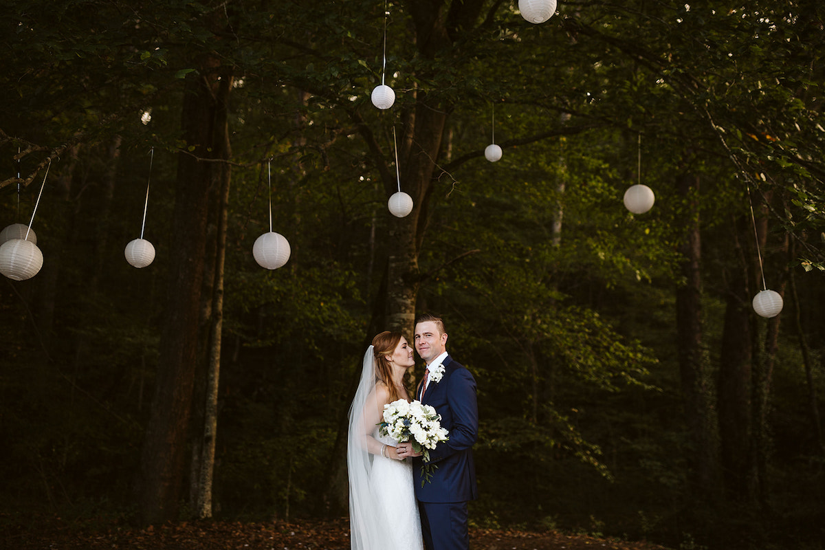 bride and groom stand beneath tall green trees hung with white paper lanterns