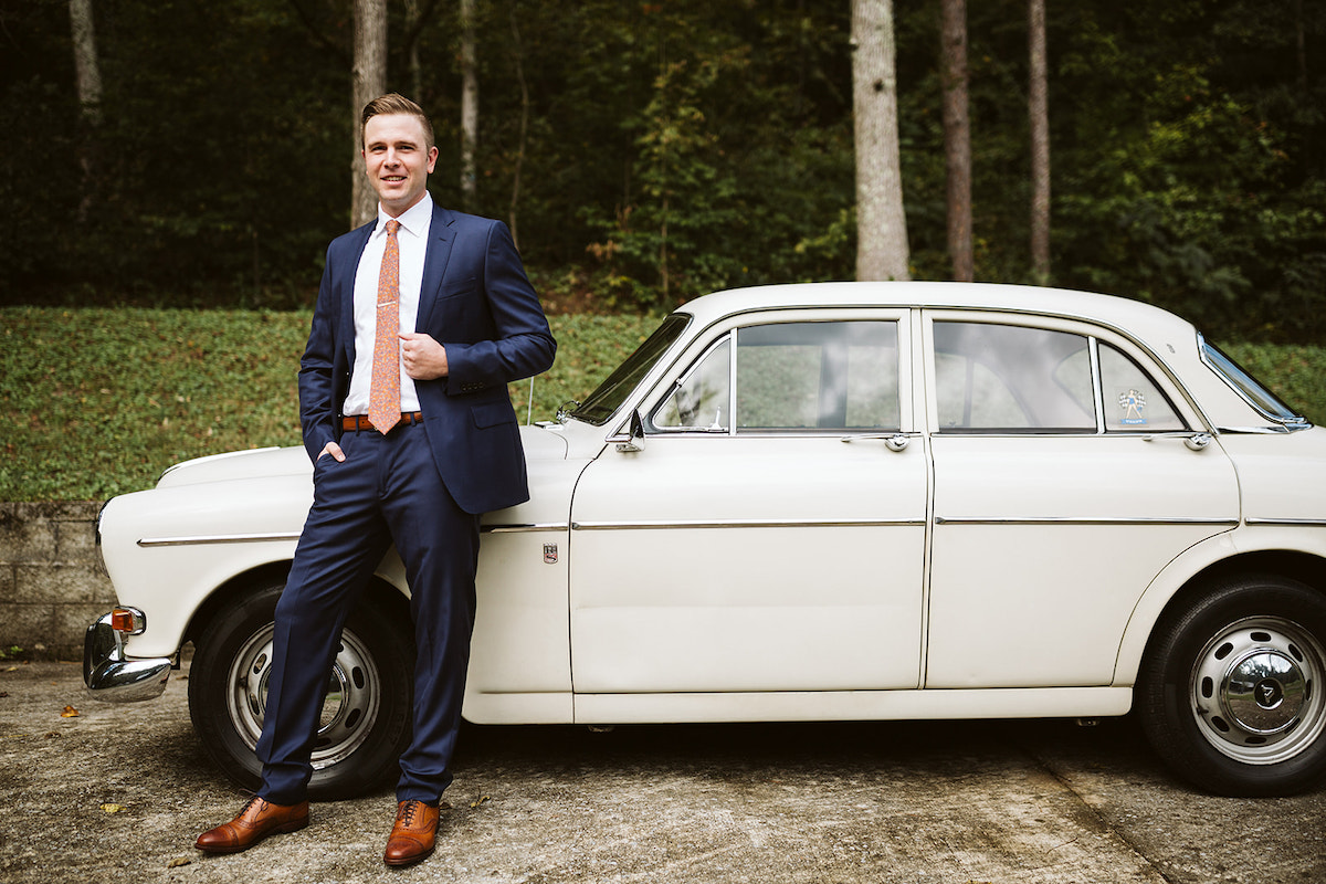 groom stands next to antique white Volvo, his hand holds onto his suit coat