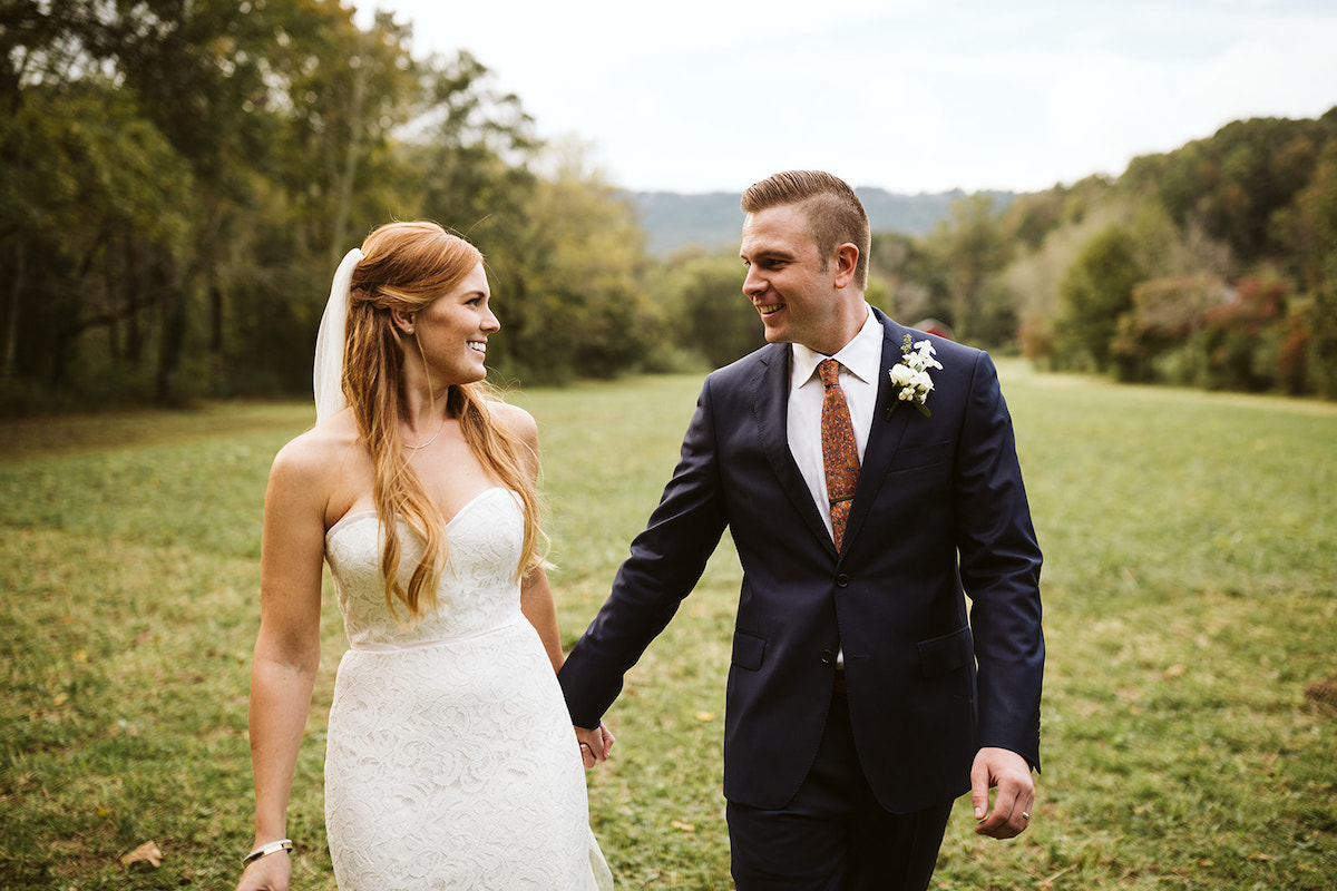 bride and groom hold hands and smile at each other as they walk through a grassy field lined with trees