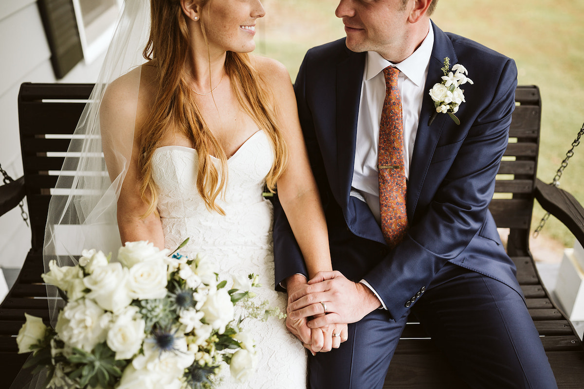 bride and groom hold hands as they sit together on a wooden porch swing