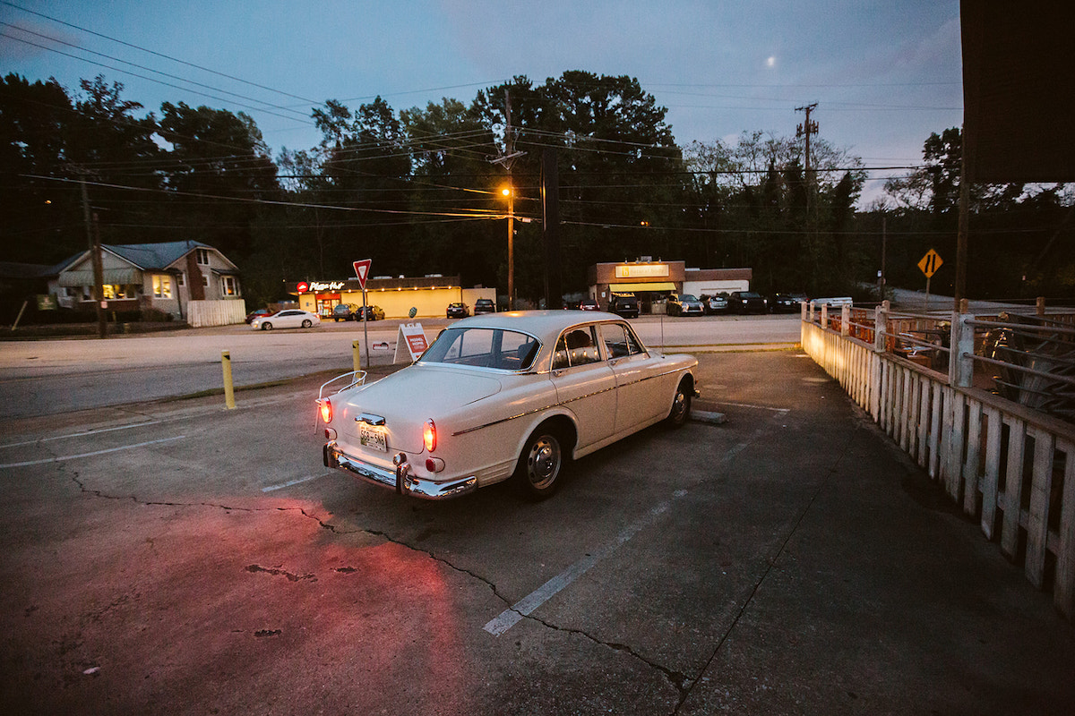 Antique white Volvo parks near a low white picket fence in the dusk light.