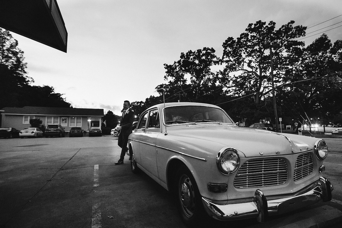 groom walks around the back of a white antique Volvo in a parking lot