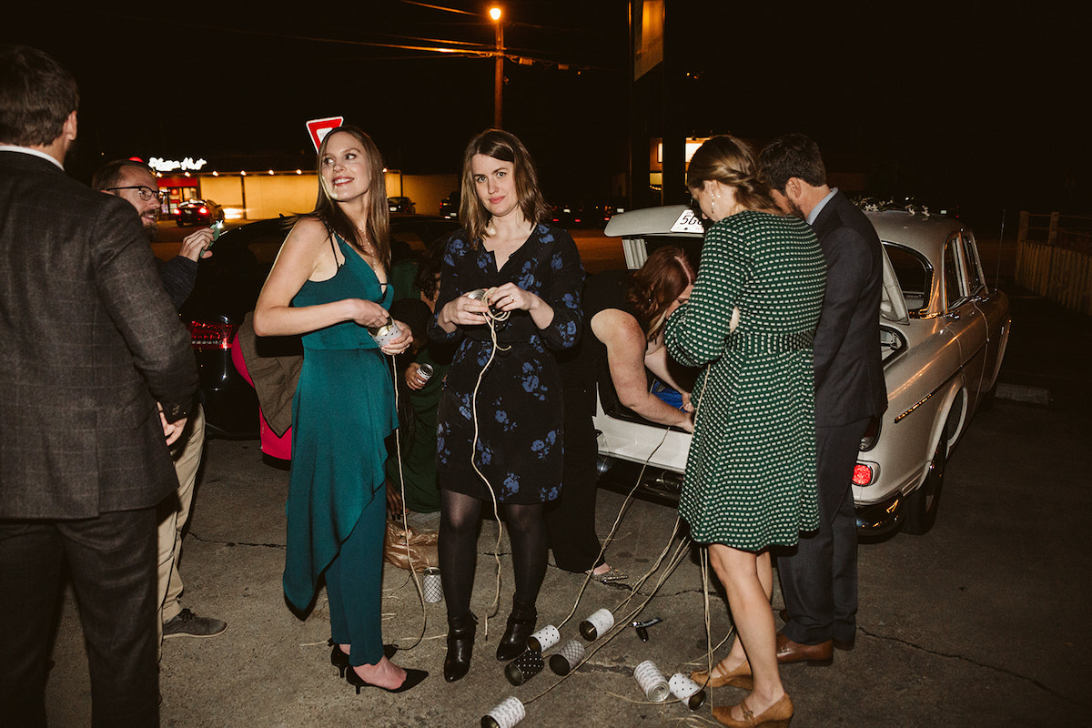 Wedding guests tie metal cans to the bumper of a white vintage automobile