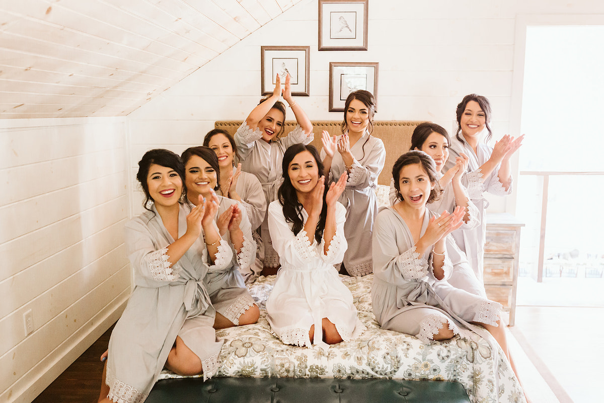 bride kneels on a bed in a white robe while bridesmaids kneels around her in gray robes