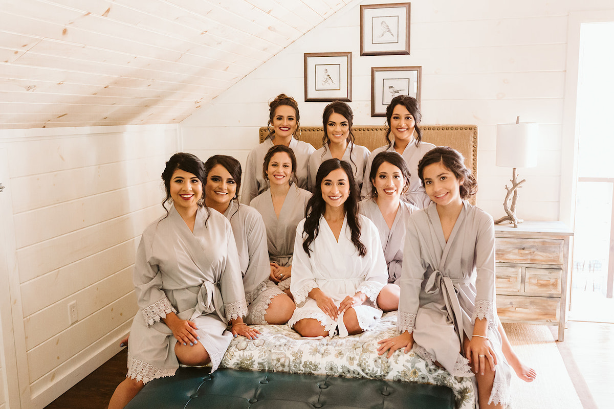 bride kneels on a bed in a white robe while bridesmaids kneels around her in gray robes