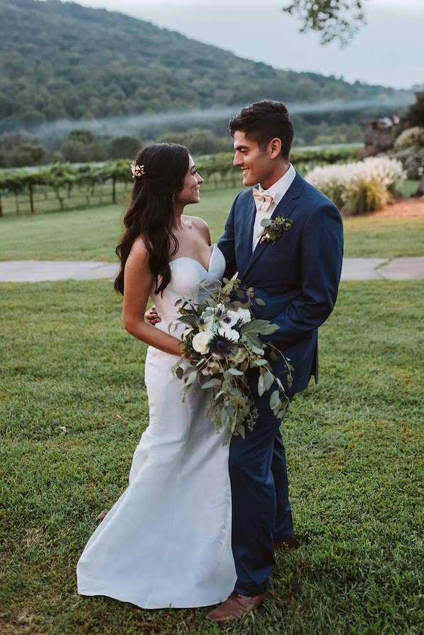 bride and groom stand in green lawn smiling at each other with rows of grape vines behind them