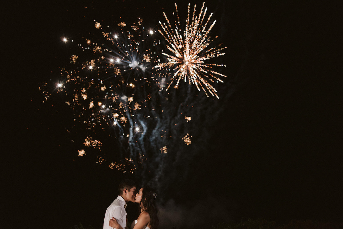 bride and groom kiss as fireworks explode in the dark sky overhead