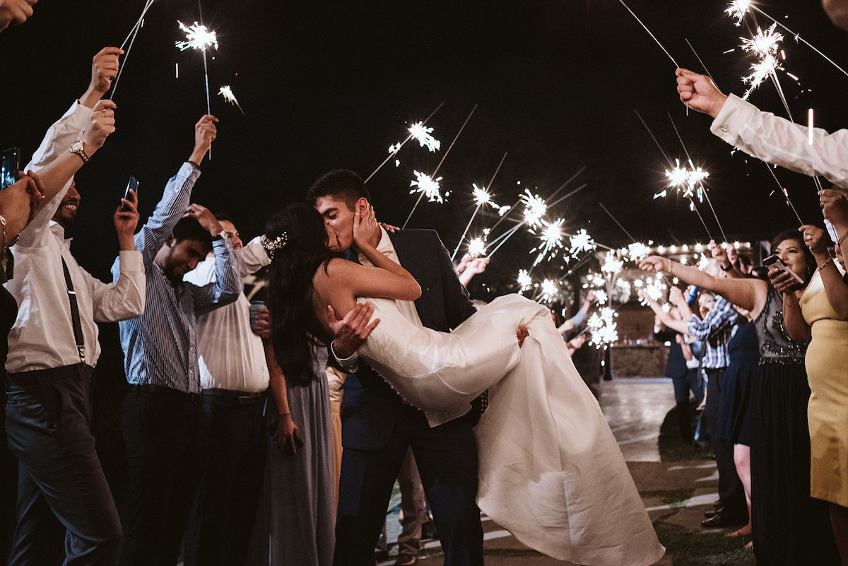 groom cradles bride in his arms and kisses her as their friends hold sparklers overhead