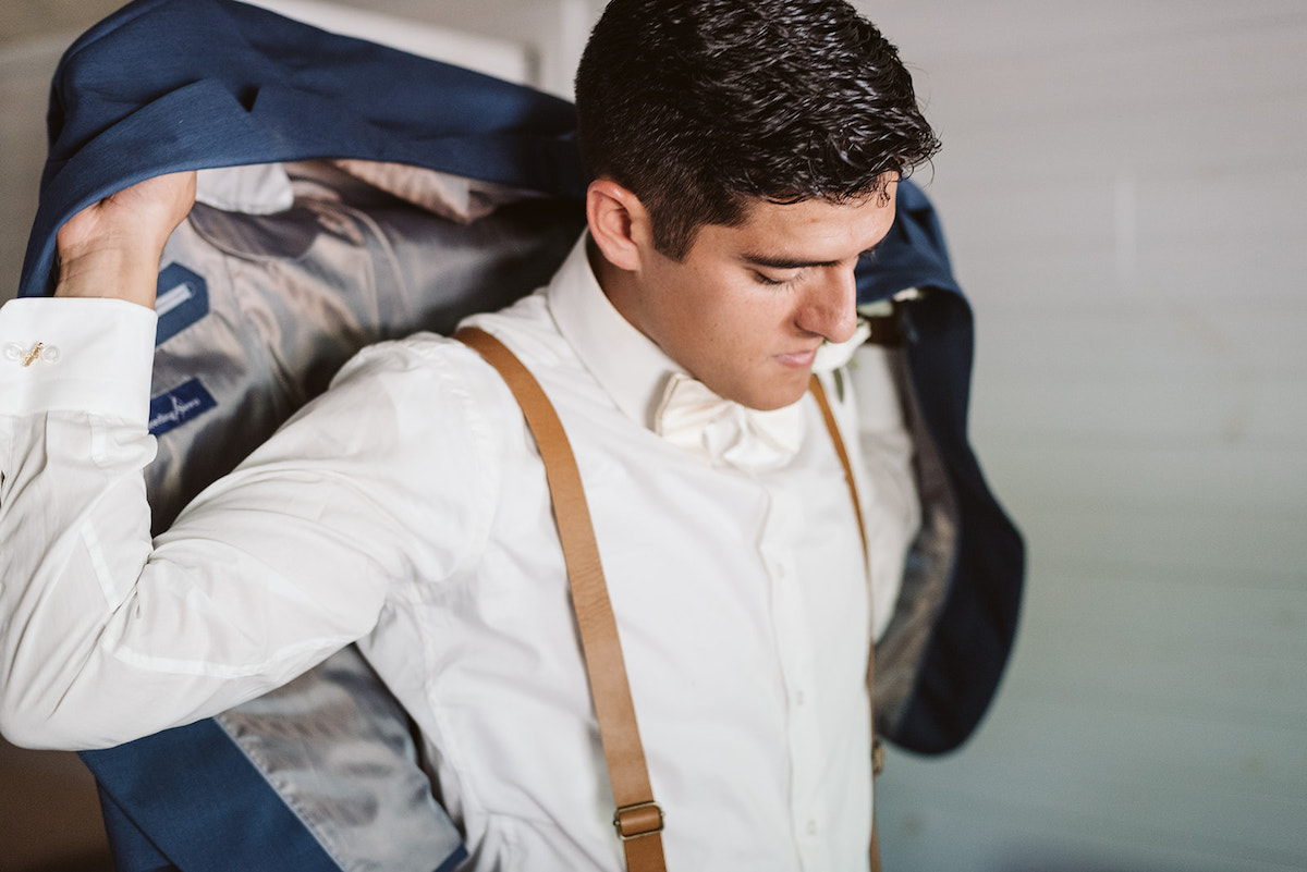 groom in white shirt, white bowtie, and brown suspenders puts on cornflower blue suit jacket