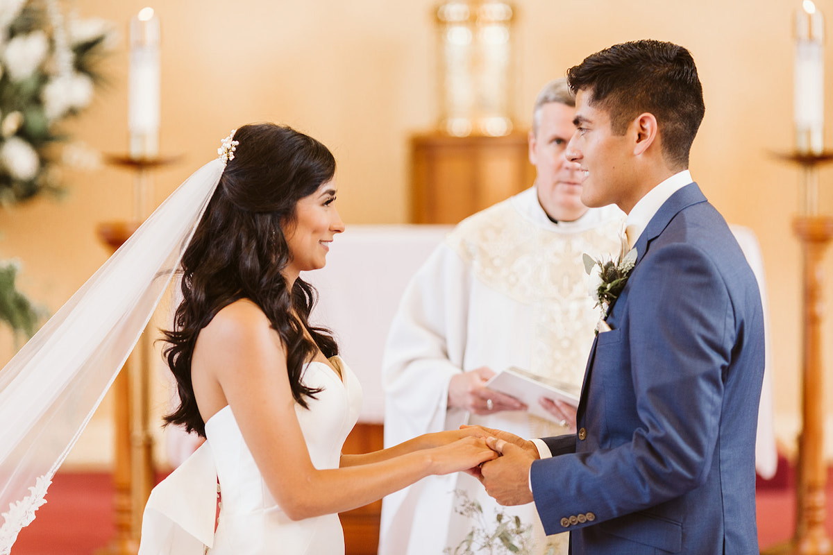 Bride and groom smile at each other and hold hands before priest