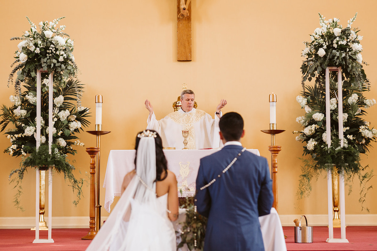 bride and groom kneel with wedding lasso rosary around their shoulders while priest raises his hands in the air