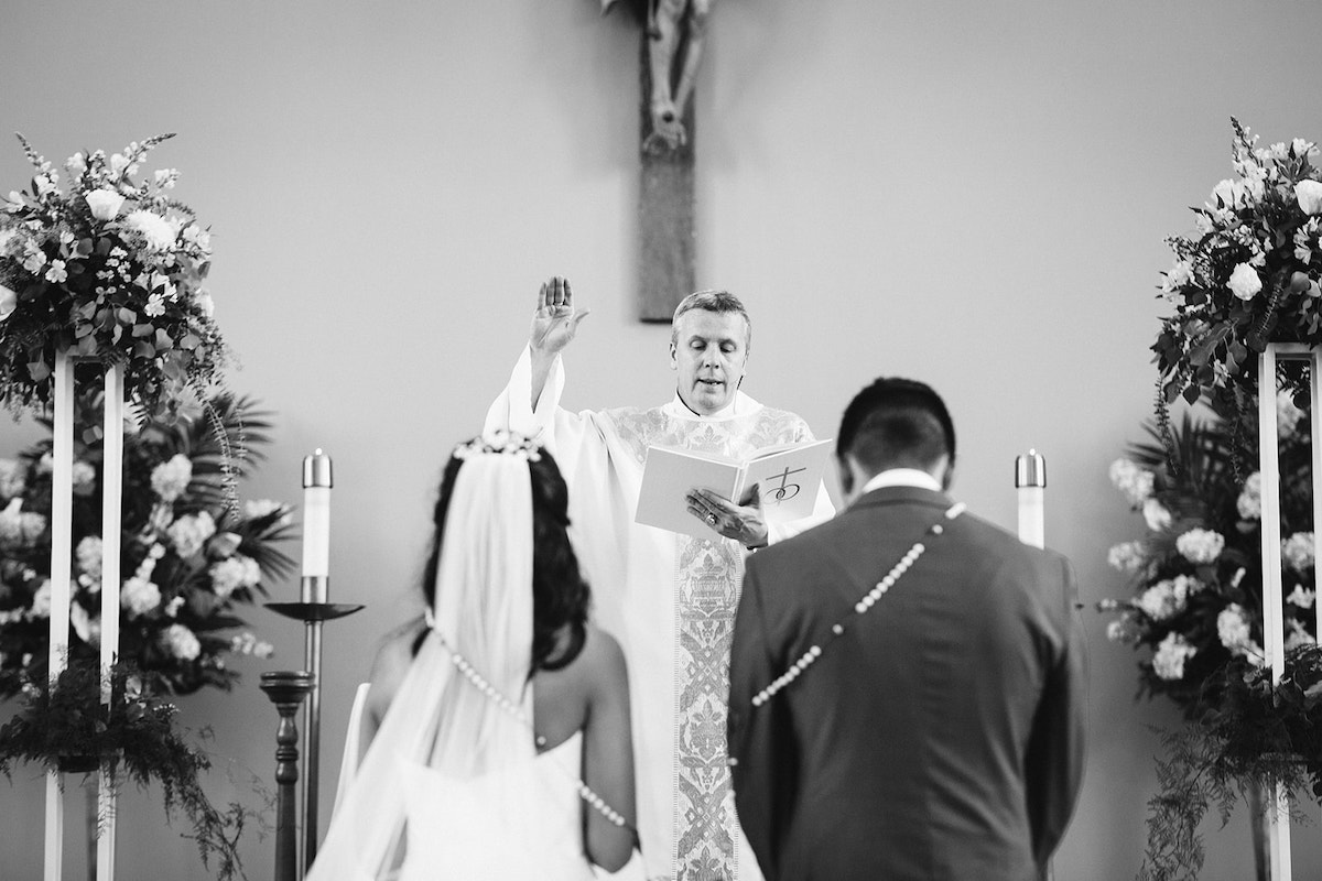 bride and groom kneel with wedding lasso rosary around their shoulders while priest raises his hands in the air