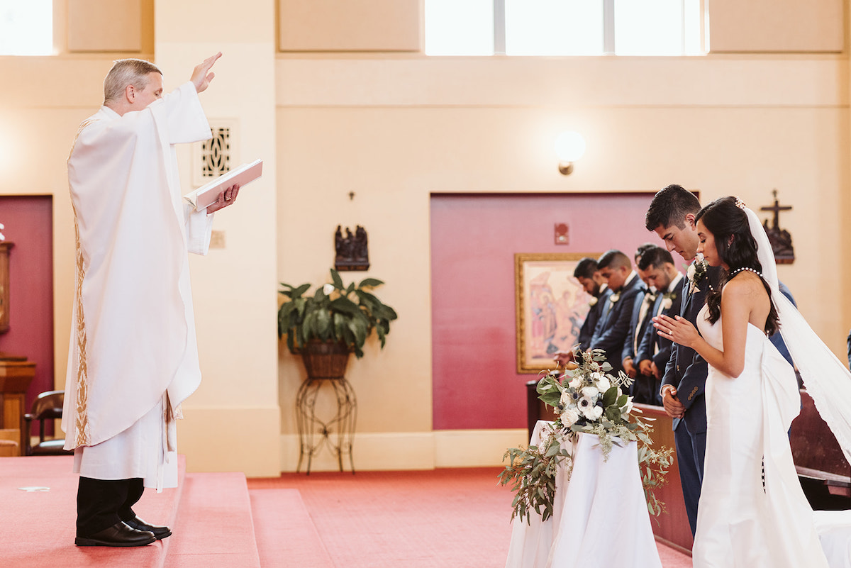 bride and groom stand in prayer with wedding lasso rosary around their shoulders while priest raises his hands in the air