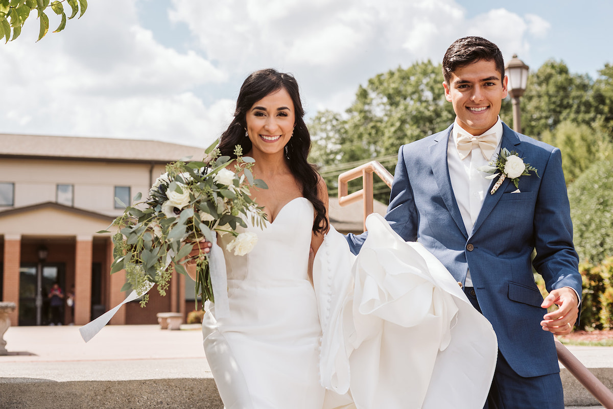 bride carries the train of her dress over one arm and bouquet in the other hand as groom helps her down the stairs