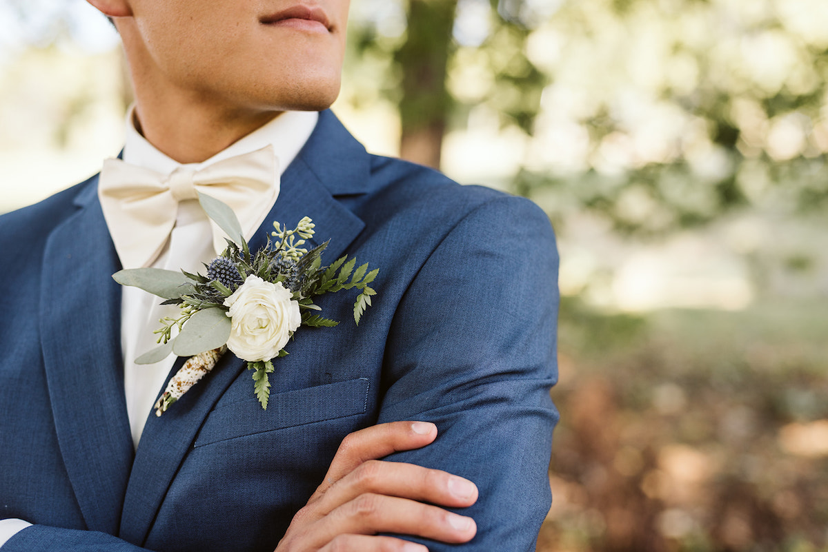 groom crosses his arms in his cornflower blue suit jacket. he wears a small boutonniere with small white rose and greens