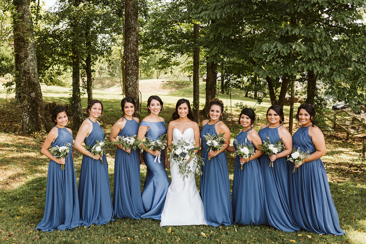 bride and bridesmaids stand in a line in the shade of large trees. they hold their bouquets of white flowers and greenery