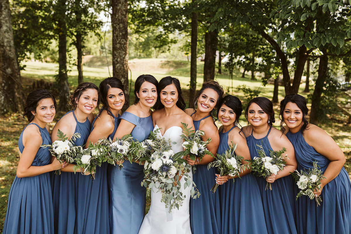 bride and bridesmaids stand in a line in the shade of large trees. they hold their bouquets of white flowers and greenery