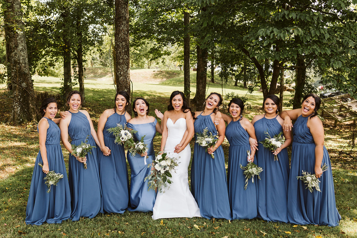 bride and bridesmaids stand in a line in the shade of large trees. they hold their bouquets of white flowers and greenery