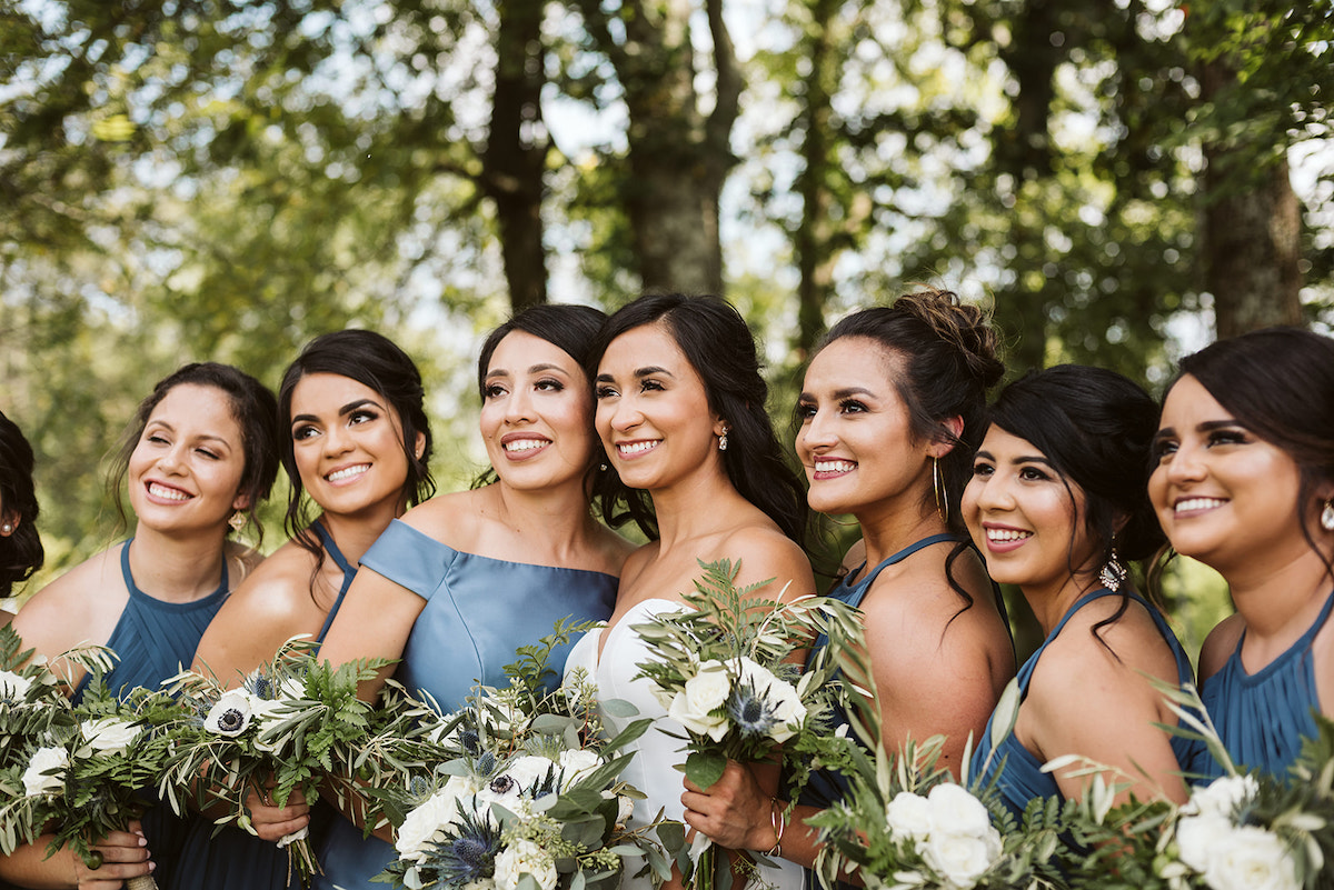 bride and bridesmaids stand in a line in the shade of large trees. they hold their bouquets of white flowers and greenery