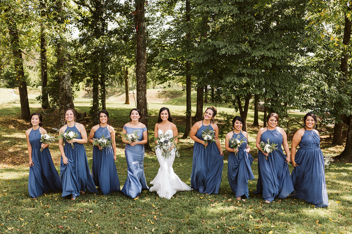 bride and bridesmaids stand in a line in the shade of large trees. they hold their bouquets of white flowers and greenery