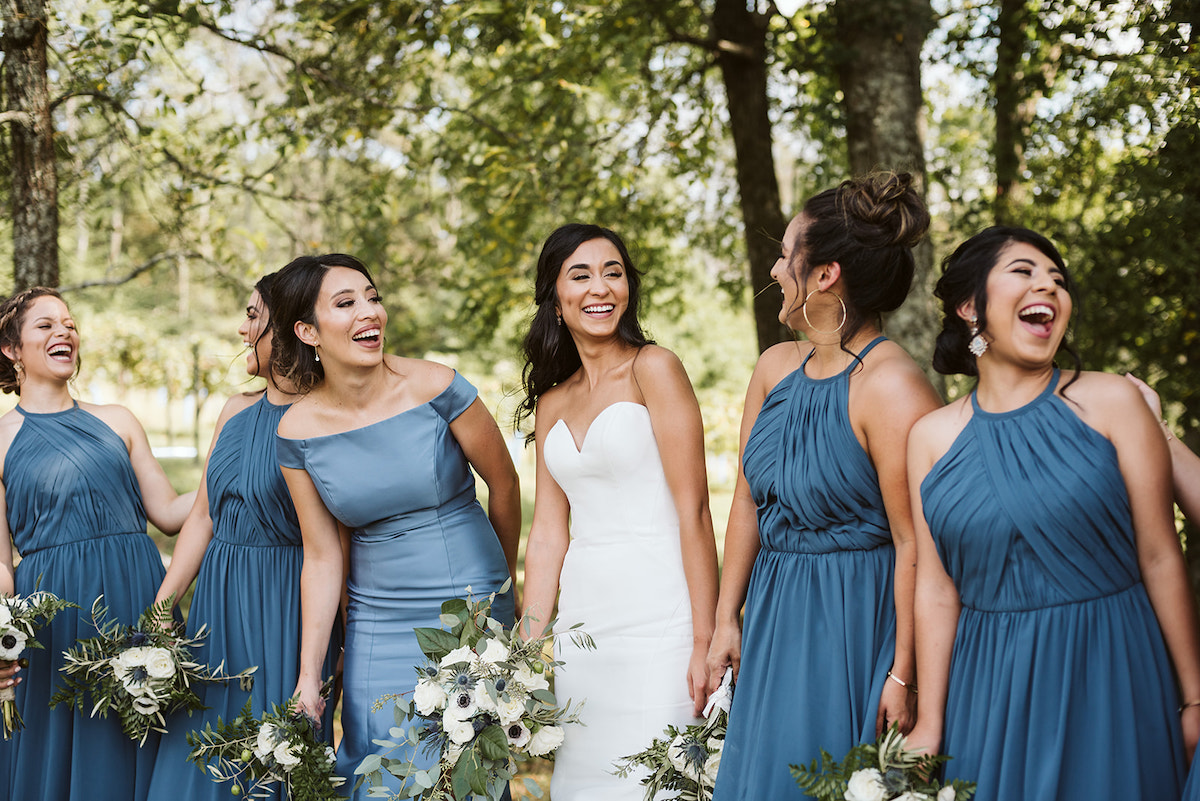 bride and bridesmaids laugh together in the shade of large trees. they hold their bouquets of white flowers and greenery