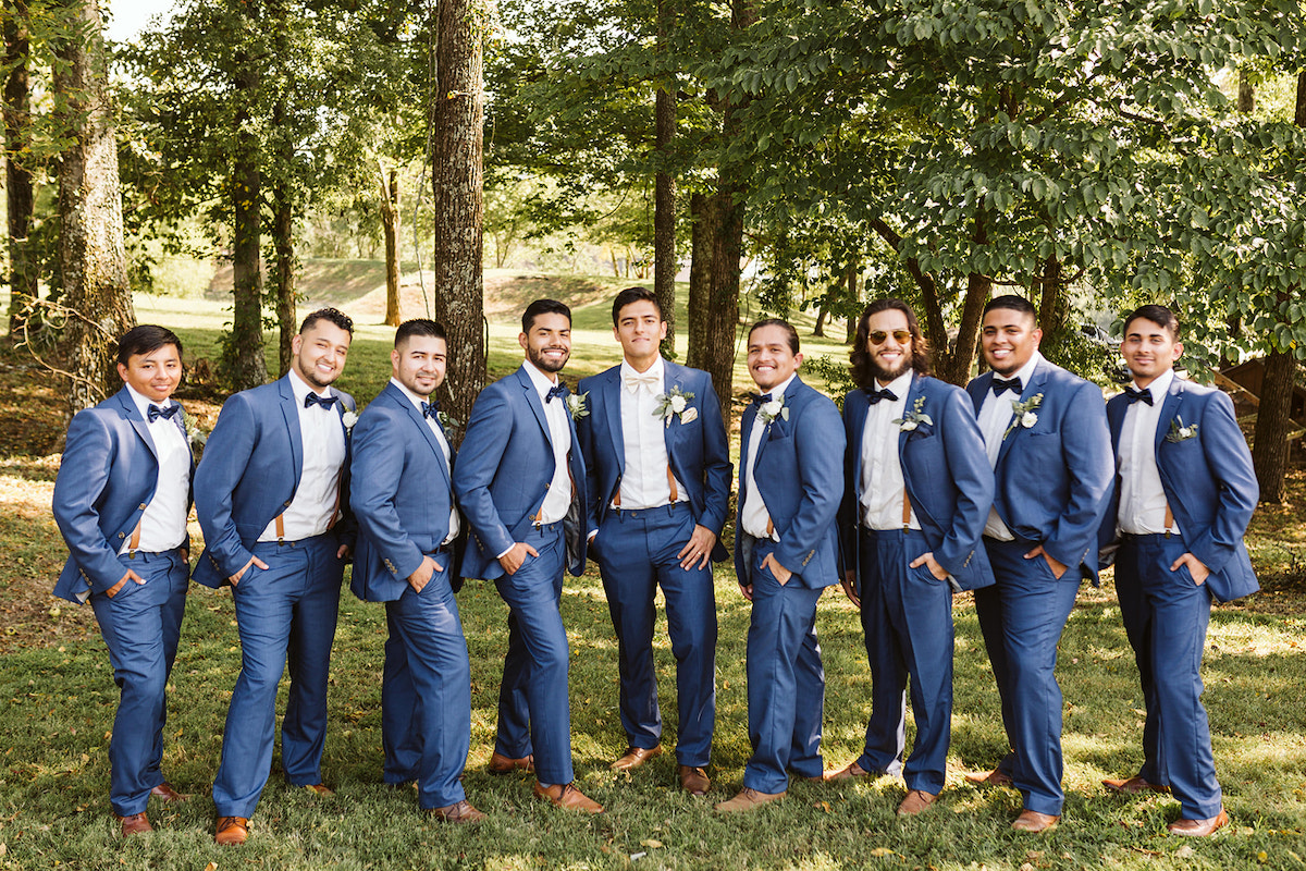 groomsmen and groom stand in a line in the shade of large trees with their hands in their pockets
