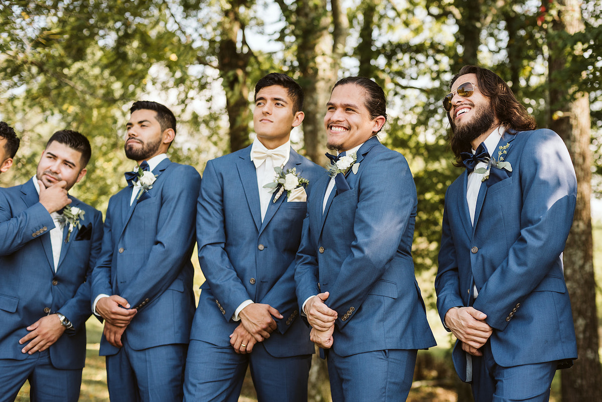 groomsmen and groom stand in a line in the shade of large trees