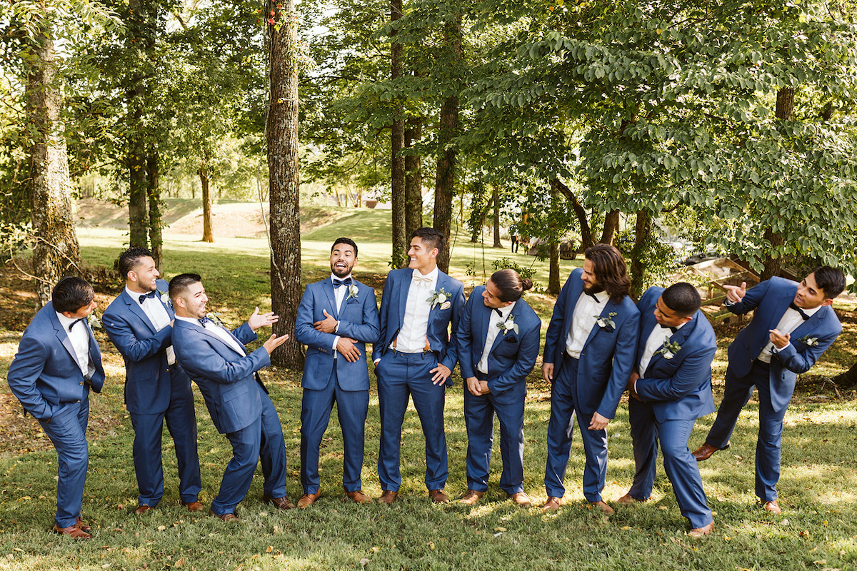 groomsmen and groom stand in a line in the shade of large trees. they joke with each other and laugh.
