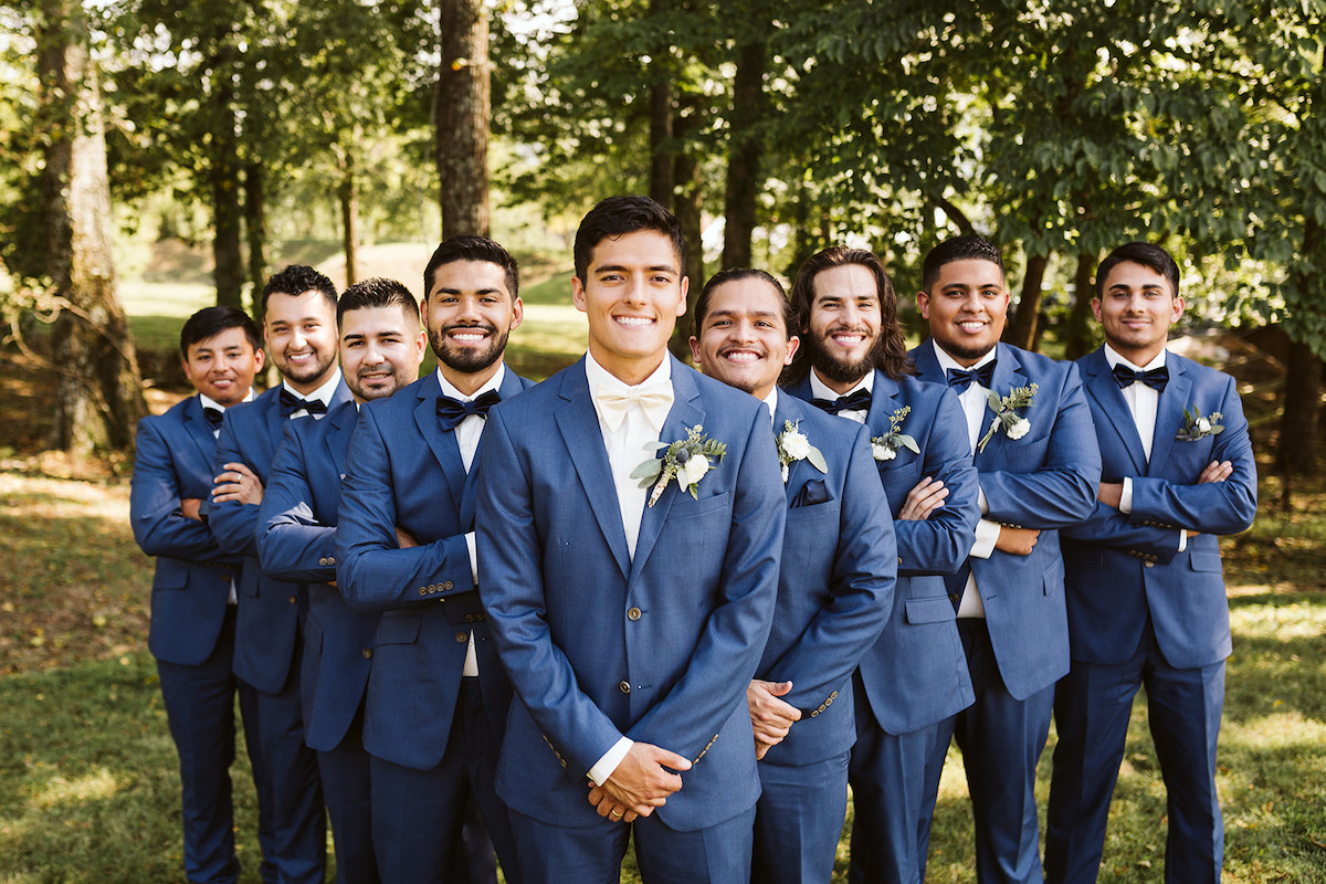 groomsmen and groom stand in the shade of large trees.