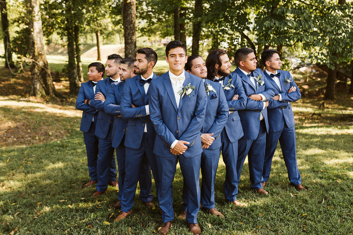 groomsmen and groom stand in the shade of large trees.