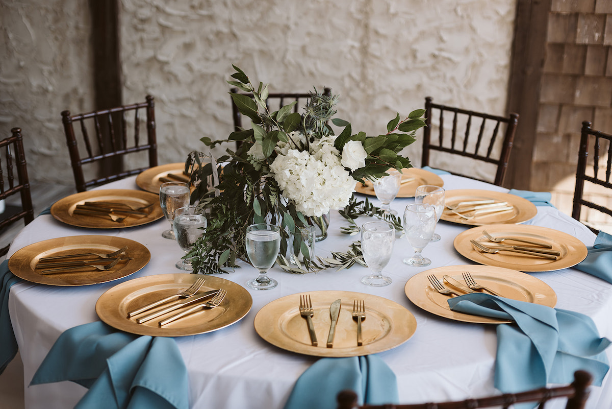 table set with gold chargers, silverware, blue cloth napkins, and white flowers and greenery