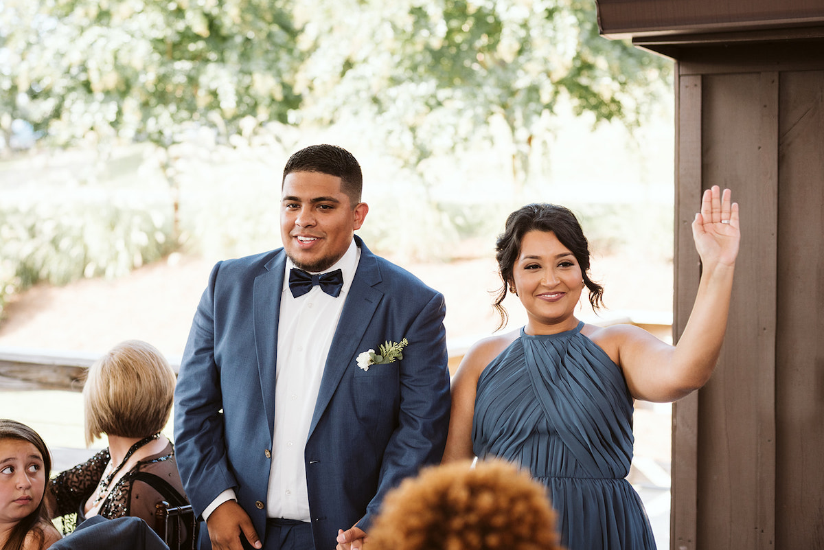bridesmaid and groomsman in cornflower blue dress and suit wave to guests