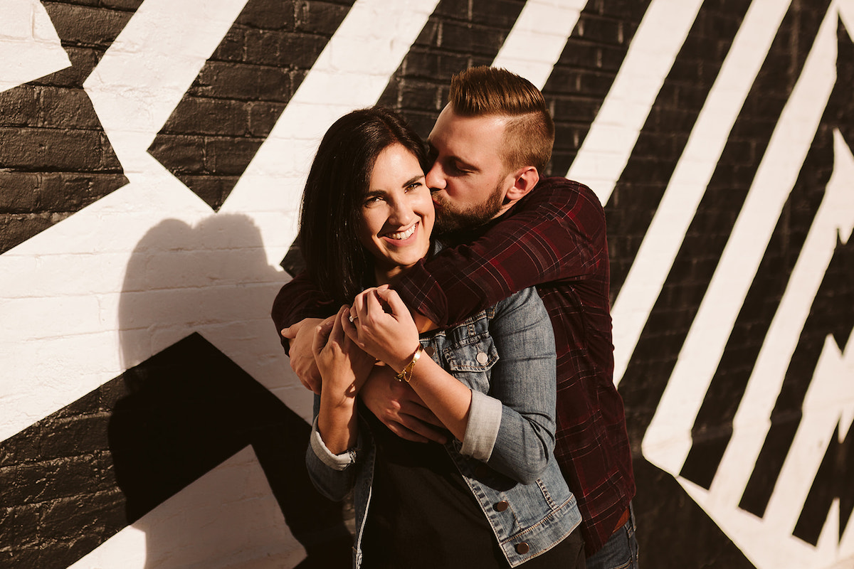 Man stands behind woman and hugs her over her shoulders and kisses her cheek. She holds his arm against her.