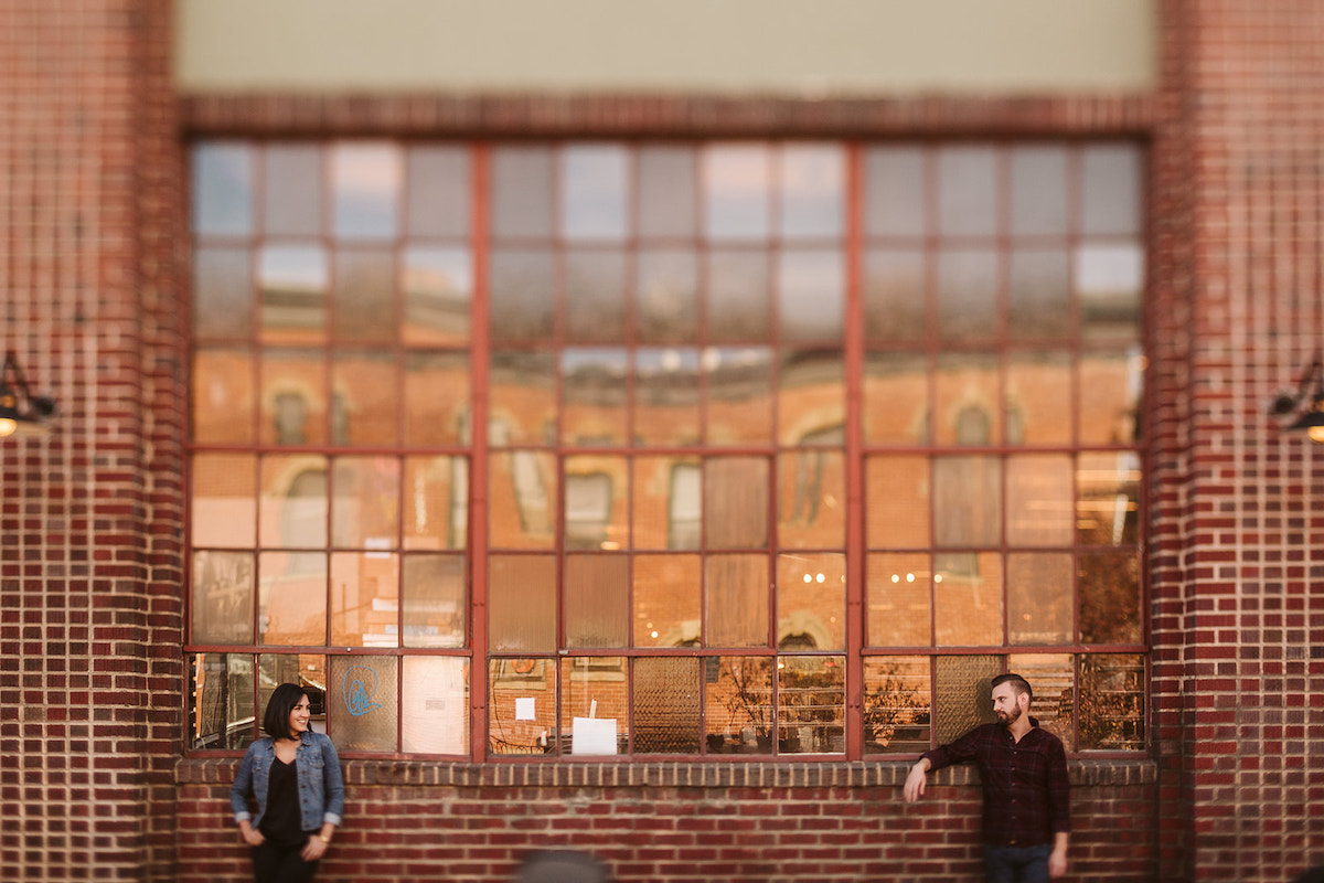 man and woman stand looking at each other against tall metal-framed windows and brick building