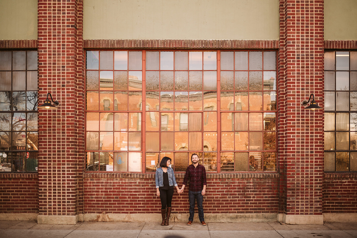 man and woman holding hands in front of tall metal-framed windows and brick building for Denver RiNo engagement photos
